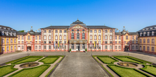 Schloss Bruchsal - Hauptbau - Von Außen - Von Osten / Hofseite - Blick vom Balkon des Torwachtgebäudes auf den Ehrenhof, den Hauptbau / das Corps de Logis (Mitte), die Verbindungsbauten (links und rechts) sowie den Kirchenflügel / die Hofkirche / St. Damian und Hugo (ganz links) und den Kammerflügel / das Amtsgericht (ganz rechts) (aufgenommen im Mai 2018, am Vormittag)