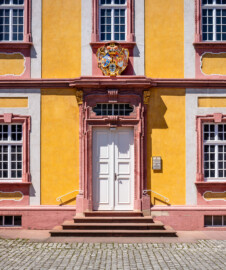 Schloss Bruchsal - Kirchenflügel - Von Osten / Hofseite - Östliche Kopfseite des Kirchenflügels (mit dem Portal zu den ehemaligen Verwaltungsräumen und dem darüber liegenden Prunkwappen von Kardinal Graf Damian Hugo von Schönborn) (aufgenommen im Mai 2018, am frühen Nachmittag)