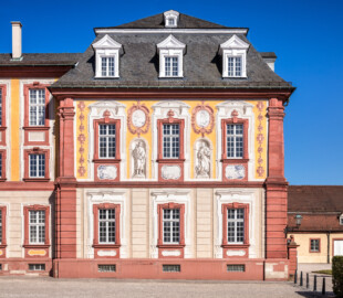 Schloss Bruchsal - Kirchenflügel / Hofkirche - Von Westen - Blick vom Kavaliersbau auf die Westfassade der Hofkirche / des Kirchenflügels mit den Heiligenfiguren von St. Damian und St. Hugo (gemalt von Walter Maschke), fünf Landschaftgrisaillen und der illusionistischen Fassadenmalerei (ursprünglich gemalt von Giovanni Francesco Marchini, zwischen 1732 und 1737) (aufgenommen im Juli 2018, am späten Nachmittag)