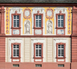Schloss Bruchsal - Kirchenflügel / Hofkirche / St. Damian und Hugo - Von aussen - Von Westen - Blick vom Kavaliersbau auf die Westfassade der Hofkirche / des Kirchenflügels mit den Heiligenfiguren von St. Damian und St. Hugo (gemalt von Walter Maschke), fünf Landschaftgrisaillen und der illusionistischen Fassadenmalerei (ursprünglich gemalt von Giovanni Francesco Marchini, zwischen 1732 und 1737) (aufgenommen im August 2018, am späten Nachmittag)