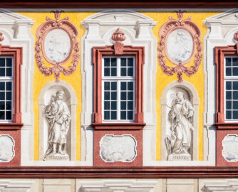 Schloss Bruchsal - Kirchenflügel / Hofkirche / St. Damian und Hugo - Von Westen - Blick vom Kavaliersbau auf den oberen Teil der Westfassade der Hofkirche mit den Heiligenfiguren von St. Damian und St. Hugo (gemalt von Walter Maschke), fünf Landschaftgrisaillen und der illusionistischen Fassadenmalerei (ursprünglich gemalt von Giovanni Francesco Marchini, zwischen 1732 und 1737) (aufgenommen im September 2018, am späten Nachmittag)