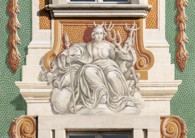 Schloss Bruchsal - Südliche Orangerie / Kavaliersbau - Von Außen - Von Westen - Blick auf die 14. (von Norden/von links gezählt) Allegorie bzw. Figur: vermutlich "Diana" (Göttin der Jagd, mit Hirsch und Lanze) (ursprünglich gemalt von Giovanni Francesco Marchini, zwischen 1732 und 1737, restauriert von Josef Mariano Kitschker, zwischen 1900 und 1910, erneut restauriert von Klaus Siller (Fassadenmalerei) und  Walter Maschke (Allegorien), 1976) (aufgenommen im September 2018, am späten Nachmittag)
