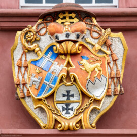 Schloss Bruchsal - Kirchenflügel / Hofkirche / St. Damian und Hugo - Von Osten / Hofseite - Prunkwappen von Kardinal Graf Damian Hugo von Schönborn über dem Ostportal - Wappenschild links: je zwei Felder für das Hochstift Speyer und die Propstei Weißenburg, Wappenschild unten: Deutschordens-Würden (Landkomtur des Deutschen Ordens in Alden-Biesen (oben) und  Marburg (unten)), Wappenschild rechts: Familien- / Stammwappen von Schönborn (aufgenommen im Oktober 2018, am frühen Nachmittag)
