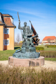 Waghäusel - Eremitage - Von Außen - Von Süden - Blick auf das Freiheitsdenkmal zum Gedenken an die Schlacht bei Waghäusel 1849 (Senkrechte Inschrift: "Den Wegbereitern der Demokratie - 21. Juni 1999"; schräge Inschrift: "In dankbarer Erinnerung an den Einsatz vieler Männer und Frauen für Freiheit und Menschenrechte in der Badischen Revolution von 1848-1849 wurde dieses Denkmal von der Stadt Waghäusel zum 150. Jahrestag der Schlacht von Waghäusel und des Gefechtes von Wiesental errichtet"; Inschrift in der Fahne: "Fond. Veneta d'Arte Verona Italia") (Skulptur aus Bronze, geschaffen 1999 von dem Speyerer Künstler Franz-Werner Müller-Steinfurt (1952-2017)) (aufgenommen im März 2019, am späten Vormittag)