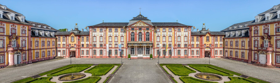 Schloss Bruchsal - Hauptbau / Corps de Logis - Von Außen / Von Osten / Hofseite - Blick vom Balkon des Torwachtgebäudes auf den Ehrenhof, den Hauptbau / das Corps de Logis (Mitte), die Verbindungsbauten (links und rechts) sowie den Kirchenflügel / die Hofkirche / St. Damian und Hugo (ganz links) und den Kammerflügel / das Amtsgericht (ganz rechts) (aufgenommen im Juni 2019, am späten Vormittag)