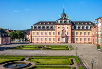 Schloss Bruchsal - Kirchenflügel / Hofkirche / St. Damian und Hugo - Von Norden / Hofseite - Blick vom Balkon des Kammerflügels / des Kammermusiksaals auf den Ehrenhof und den Kirchenflügel, dahinter der Kirchturm mit den Fronleichnamsfahnen, links das Hofzahlamt /  das Schlosscafé und rechts der Hauptbau / das Corps de Logis (aufgenommen im Juni 2019, am Vormittag)