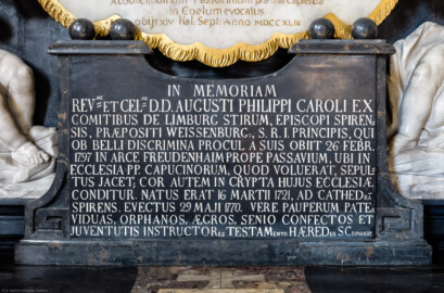Bruchsal - Peterskirche / St. Peter - Westlicher Chorraum - Doppelgrabdenkmal für Damian Hugo Philipp von Schönborn-Buchheim und für August von Limburg-Stirum - Von Innen / Von Nordost - Blick auf die Gedenktafel für August von Limburg-Stirum am unteren Teil des Grabdenkmals; die Inschrift lautet auf Deutsch: "Zum Gedenken an den verehrungswürdigsten und hochberühmten Herrn, Herrn August Philipp Karls aus dem Geschlecht der Grafen von Limburg-Stirum, Bischof von Speyer, Probst von Weißenburg, Fürst des Heiligen Römischen Reiches, der wegen Kriegswirren fern von den Seinen am 26. Februar 1797 in Burg Freudenhain in der Nähe von Passau starb, wo er in der Kirche der Kapuziner, was er gewünscht hatte, begraben liegt; sein Herz aber wird in der Krypta dieser Kirche verwahrt. Geboren war er am 16. März 1721, auf den Bischofsstuhl von Speyer erhoben am 29. Mai 1770. Wahrhaft ein Vater der Armen hatte er Witwen, Waisen, Kranke, vom Alter Geschwächte und die Erzieher der Jugend in seinem Testament schriftlich als Erben eingesetzt."  (Epitaph entworfen von Johann Seitz, gefertigt von Stephan Strahl, vorgelagerter Marmoraltar und dazugehörende Plastiken gefertigt von Joachim Günther) (aufgenommen im September 2019, um die Mittagszeit)