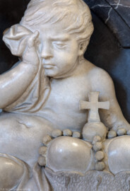Bruchsal - Peterskirche / St. Peter - Westlicher Chorraum - Doppelgrabdenkmal für Damian Hugo Philipp von Schönborn-Buchheim und für August von Limburg-Stirum - Von Nordost - Blick auf das trauernde Gesicht einer Knabenfigur / eines Putto, unten der Bischofshut (weiter unten, rechts am Grabdenkmal; gefertigt von Joachim Günther) (aufgenommen im September 2019, am späten Nachmittag)