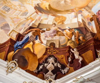 Schloss Bruchsal - Hauptbau / Corps de Logis - Beletage - Fürstensaal - Decke, untere Zone, Nordostecke - Ausschnitt aus der nordöstlichen äußeren Zone - Darstellung der Salzgewinnung, ein wichtiger Wirtschaftszweig Bruchsals - Mittig oben (Frau mit Schlüssel in der Hand): Darstellung der Erdgöttin Cybele mit den Attributen der Erde (Mauerkrone, Löwe und Früchtekorb); aus ihrer Brust fließt Salzwasser, das auf dem Herd verdampft; links (Mann mit Hammer): Darstellung des Vulcanus, des Feuer- und Schmiedegotts; rechts (Männer bei der Salzabfüllung): Salz wird in einen Sack abgefüllt, der Sack trägt das alchemistische Zeichen für Salz (Deckenfresko urspünglich gemalt 1751 von Johannes Zick, originalgetreu rekonstruiert 1966 bis 1969 von Wolfram Köberl) (aufgenommen im Dezember 2019, am späten Vormittag)