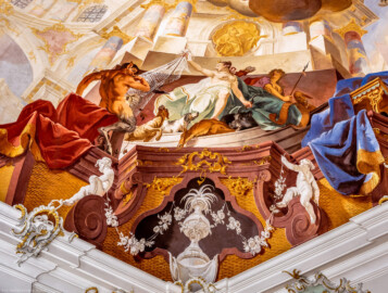 Schloss Bruchsal - Hauptbau / Corps de Logis - Beletage - Fürstensaal - Decke, untere Zone, Südwestecke - Ausschnitt aus der südwestlichen äußeren Zone - Darstellung der Jagd, ein wichtiger Wirtschaftszweig Bruchsals - Mittig (Frau mit Bogen und Köcher): Darstellung der griechischen / römischen Jagdgöttin Artemis / Diana, auf dem Kopf eine Mondsichel (als Göttin des Mondes); links (mannartige Figur); ein Faun, halb Mensch, halb Bock, ein Waldwesen, Begleiter der Jagdgöttin (Deckenfresko urspünglich gemalt 1751 von Johannes Zick, originalgetreu rekonstruiert 1966 bis 1969 von Wolfram Köberl) (aufgenommen im Januar 2020, um die Mittagszeit)