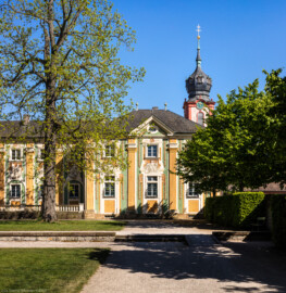 Schloss Bruchsal - Südliche Orangerie / Kavaliersbau - Von Außen / Von Westen - Blick von der südlichsten Ost-West-Achse auf die Westfassade der Orangerie, im Hintergrund der Kirchturm der Hofkirche / von St. Damian und Hugo (Fassade der Orangerie ursprünglich gemalt von Giovanni Francesco Marchini, zwischen 1732 und 1737, restauriert von Josef Mariano Kitschker, zwischen 1900 und 1910, erneut restauriert von Klaus Siller (Fassadenmalerei) und  Walter Maschke (Allegorien), 1976) (aufgenommen im April 2020, am späten Nachmittag)