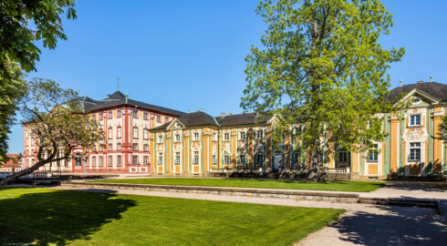 Schloss Bruchsal - Südliche Orangerie / Kavaliersbau, Westseite - Von Südwest - Blick von der südlichsten Ost-West-Achse auf die Westfassade der Orangerie (rechts) sowie auf die Südwestseite des Hauptbaus / des Corps de Logis (links) (Fassade der Orangerie ursprünglich gemalt von Giovanni Francesco Marchini, zwischen 1732 und 1737, restauriert von Josef Mariano Kitschker, zwischen 1900 und 1910, erneut restauriert von Klaus Siller (Fassadenmalerei) und  Walter Maschke (Allegorien), 1976) (aufgenommen im Mai 2020, am späten Nachmittag)