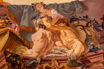 Schloss Bruchsal - Hauptbau / Corps de Logis - Beletage - Fürstensaal - Decke, äußere Zone, Südostecke - Ausschnitt aus der südöstlichen äußeren Zone - Ausschnitt aus der Darstellung des Weinbaus, ein wichtiger Wirtschaftszweig Bruchsals - Mittig oben (Mann/Frau mit Weinlaubkrone und Korb mit Weintrauben): Darstellung des griechischen / römischen Weingottes Dionysos / Bacchus; mittig unten (mannartige Figuren): Darstellung von Satyrn, mit Bocks-Beinen und -Ohren; rechts (Ziegenbock): Darstellung der vollendeten Umwandlung vom Menschen zum Bock bei übermäßigem Konsum von Wein (Deckenfresko urspünglich gemalt 1751 von Johannes Zick, originalgetreu rekonstruiert 1966 bis 1969 von Wolfram Köberl) (aufgenommen im Mai 2020, am späten Vormittag) Schloss Bruchsal - Hauptbau / Corps de Logis - Beletage - Fürstensaal - Decke, äußere Zone, Südostecke - Ausschnitt aus der südöstlichen äußeren Zone - Ausschnitt aus der Darstellung des Weinbaus, ein wichtiger Wirtschaftszweig Bruchsals - Mittig oben (Mann/Frau mit Weinlaubkrone und Korb mit Weintrauben): Darstellung des griechischen / römischen Weingottes Dionysos / Bacchus; mittig unten (mannartige Figuren): Darstellung von Satyrn, mit Bocks-Beinen und -Ohren; rechts (Ziegenbock): Darstellung der vollendeten Umwandlung vom Menschen zum Bock bei übermäßigem Konsum von Wein (Deckenfresko urspünglich gemalt 1751 von Johannes Zick, originalgetreu rekonstruiert 1966 bis 1969 von Wolfram Köberl) (aufgenommen im Mai 2020, am späten Vormittag)