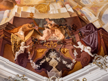 Schloss Bruchsal - Hauptbau / Corps de Logis - Beletage - Fürstensaal - Decke, untere Zone, Südostecke - Ausschnitt aus der südöstlichen äußeren Zone - Darstellung des Weinbaus, ein wichtiger Wirtschaftszweig Bruchsals - Mittig oben (Mann/Frau mit Weinlaubkrone und Korb mit Weintrauben): Darstellung des griechischen / römischen Weingottes Dionysos / Bacchus; mittig unten (mannartige Figuren): Darstellung von Satyrn, mit Bocks-Beinen und -Ohren; links (Mann mit Weinglas); Bacchant, Teilnehmer der Zeche; rechts (Ziegenbock): Darstellung der vollendeten Umwandlung vom  Menschen zum Bock bei übermäßigem Konsum von Wein (Deckenfresko urspünglich gemalt 1751 von Johannes Zick, originalgetreu rekonstruiert 1966 bis 1969 von Wolfram Köberl) (aufgenommen im Mai 2020, um die Mittagszeit)