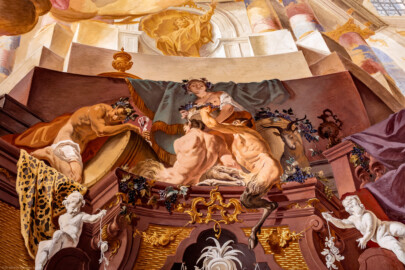 Schloss Bruchsal - Hauptbau / Corps de Logis - Beletage - Fürstensaal - Decke, untere Zone, Südostecke - Ausschnitt aus der südöstlichen äußeren Zone - Darstellung des Weinbaus, ein wichtiger Wirtschaftszweig Bruchsals - Mittig oben (Mann/Frau mit Weinlaubkrone und Korb mit Weintrauben): Darstellung des griechischen / römischen Weingottes Dionysos / Bacchus; mittig unten (mannartige Figuren): Darstellung von Satyrn, mit Bocks-Beinen und -Ohren; links (Mann mit Weinglas); Bacchant, Teilnehmer der Zeche; rechts (Ziegenbock): Darstellung der vollendeten Umwandlung vom  Menschen zum Bock bei übermäßigem Konsum von Wein (Deckenfresko urspünglich gemalt 1751 von Johannes Zick, originalgetreu rekonstruiert 1966 bis 1969 von Wolfram Köberl) (aufgenommen im Mai 2020, um die Mittagszeit)