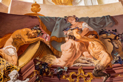 Schloss Bruchsal - Hauptbau / Corps de Logis - Beletage - Fürstensaal - Decke, untere Zone, Südostecke - Ausschnitt aus der südöstlichen äußeren Zone - Darstellung des Weinbaus, ein wichtiger Wirtschaftszweig Bruchsals - Mittig oben (Mann/Frau mit Weinlaubkrone und Korb mit Weintrauben): Darstellung des griechischen / römischen Weingottes Dionysos / Bacchus; mittig unten (mannartige Figuren): Darstellung von Satyrn, mit Bocks-Beinen und -Ohren; links (Mann mit Weinglas); Bacchant, Teilnehmer der Zeche; rechts (Ziegenbock): Darstellung der vollendeten Umwandlung vom  Menschen zum Bock bei übermäßigem Konsum von Wein (Deckenfresko urspünglich gemalt 1751 von Johannes Zick, originalgetreu rekonstruiert 1966 bis 1969 von Wolfram Köberl) (aufgenommen im Mai 2020, am frühen Nachmittag)