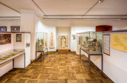 Städtisches Museum Bruchsal - Schloss Bruchsal - Hauptbau / Corps de Logis - Südlicher Raum, Bereich Mittelalter bis Neuzeit - Blick nach Norden, in der Mitte das Epitaph von Gerhard von Ehrenberg (1336-1363, Bischof von Speyer) sowie ein Plan des Bruchsaler Bergfrieds aus dem Jahr 1737, links Exponate zum ehemaligen Kapuzinerkloster Bruchsal sowie Vitrinen mit Keramik- und Glasfunden, rechts ein alter Plan und eine alte Stadtansicht von Bruchsal, eine Vitrine mit Keramikfunden aus dem Alten Schloss sowie Exponate zur ehemaligen Bischöflichen Burg Bruchsal (aufgenommen im November 2020, am späten Vormittag)