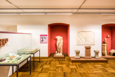 Städtisches Museum Bruchsal - Schloss Bruchsal - Hauptbau / Corps de Logis - Südlicher Raum, Bereich Römerzeit - Blick nach Norden, links Vitrinen mit diversen Fundstücken, an der Wand ein Dreigötterstein, in der Mitte eine Skulptur "Herkules", rechts davon ein Hausaltar, ein Votivstein der Vierwegegöttinnen, ein weiterer Hausaltar, ein Weihestein, ein Kopf und ein Götterstein der Göttin Minerva sowie ein Totenhaus (aufgenommen im November 2020, am frühen Nachmittag)