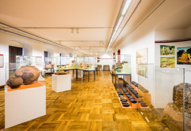 Städtisches Museum Bruchsal - Schloss Bruchsal - Hauptbau / Corps de Logis - Von innen - Südlicher Raum, nordlicher Teil, Bereiche Bronze- / Latène- / Hallstatt- / Römerzeit - Von Osten - Blick in den Raum nach Westen, links vorne Kermaikgefäße aus der Hallstattzeit, dahinter eine Getreide-Handmühle aus der Latènezeit, dahinter Vitrinen mit römischen Fundstücken, rechts ein nachgebauter Rennofen zur experimentellen Eisenverhüttung, dahinter ein Eisenbarrenfund mit Doppelpyramidenbarren, dahinter eine Vitrine mit römischen Bronzegefäßen (aufgenommen im Dezember 2020, am frühen Nachmittag)