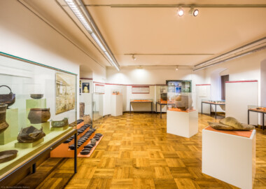 Städtisches Museum Bruchsal - Schloss Bruchsal - Hauptbau / Corps de Logis - Von innen - Südlicher Raum, nordlicher Teil, Bereiche Bronze- / Latène- / Hallstatt- / Römerzeit - Von Westen - Blick in den Raum nach Osten, links eine Vitrine mit römischen Bronzegefäßen, dahinter ein Eisenbarrenfund mit Doppelpyramidenbarren, dahinter ein nachgebauter Rennofen zur experimentellen Eisenverhüttung, in der Mitte Vitrinen mit Fundstücken aus der Hallstatt- und Latènezeit, rechts eine Getreide-Handmühle aus der Latènezeit, dahinter Kermaikgefäße aus der Hallstattzeit, ganz rechzts Vitrinen mit Fundstücken aus der Bronze- und Römerzeit (aufgenommen im Dezember 2020, am frühen Nachmittag)