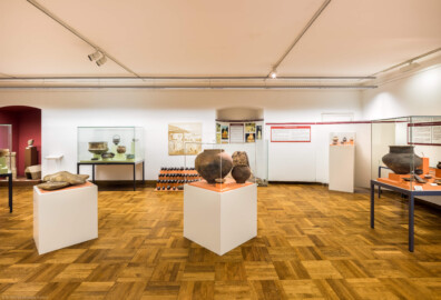 Städtisches Museum Bruchsal - Schloss Bruchsal - Hauptbau / Corps de Logis - Von innen - Südlicher Raum, Bereiche Bronze-  / Hallstatt- / Latènezeit - Von Süden - Blick auf Vitrinen und Tafeln zur Bronze-, Hallstatt- und Latènezeit; vorne links eine Getreide-Handmühle aus der Latènezeit, vorne mittig Kermaikgefäße aus der Hallstattzeit, vorne rechts Kermaikgefäße aus einem Urnengrab der Bronzezeit, hinten links an der Wand römische Bronzegefäße, rechts davon ein Eisenbarrenfund mit Doppelpyramidenbarren, rechts davon (hinter der Vitrine) ein nachgebauter Rennofen zur Eisenverhüttung und hinten rechts Kermaikgefäße aus der Latènezeit (aufgenommen im Dezember 2020, am frühen Nachmittag)