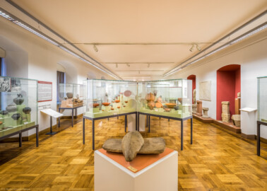 Städtisches Museum Bruchsal - Schloss Bruchsal - Hauptbau / Corps de Logis - Von innen - Südlicher Raum, Bereiche Latènezeit / Römerzeit - Von Osten - Blick nach Westen, vorne eine Getreide-Handmühle, in der Mitte Vitrinen mit römischem Geschirr, links Vitrinen mit Funden der Römer- und Merowingerzeit, rechts ein Totenhaus, ein Kopf und ein Götterstein, ein Weihestein, Hausaltare, ein Votivstein sowie eine Skulptur "Herkules" (aufgenommen im Dezember 2020, um die Mittagszeit)