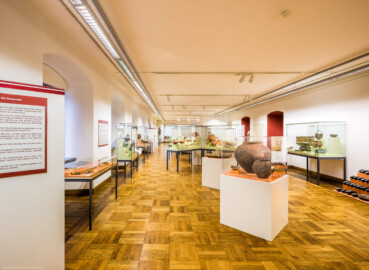 Städtisches Museum Bruchsal - Schloss Bruchsal - Hauptbau / Corps de Logis - Von innen - Südlicher Raum, südliche Hälfte, Bereiche Bronze- / Latène- / Hallstatt- / Römerzeit - Von Osten - Blick vom Eingang in den Raum nach Westen, links Vitrinen mit Fundstücken aus der Bronze- und Römerzeit, in der Mitte vorne Kermaikgefäße aus der Hallstattzeit, dahinter eine Getreide-Handmühle aus der Latènezeit, dahinter Vitrinen mit römischen Fundstücken, rechts ein Eisenbarrenfund mit Doppelpyramidenbarren, dahinter eine Vitrine mit römischen Bronzegefäßen (aufgenommen im Dezember 2020, am frühen Nachmittag)