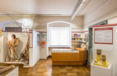Städtisches Museum Bruchsal - Schloss Bruchsal - Hauptbau / Corps de Logis - Von innen - Östlicher Raum, Bereich Jungsteinzeit / Infostand - Von Norden - Blick nach Süden auf den Infobereich, links ein Modell eines jungsteinzeitlichen Jägers, rechts eine Vitrine mit Glockenbechern und einem Dolch aus Feuerstein (aufgenommen im März 2021, am frühen Nachmittag)