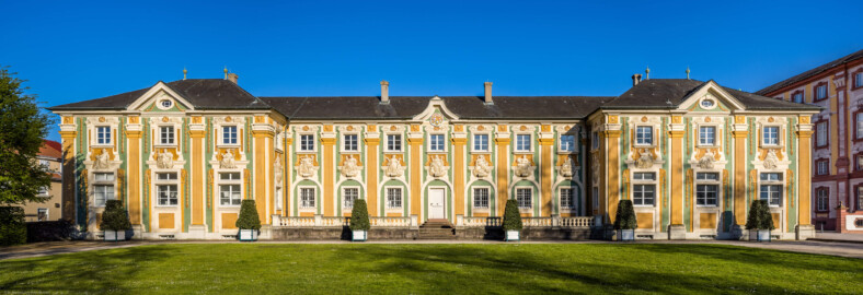 Schloss Bruchsal - Nördliche Orangerie / Nebendienstdienerbau - Von Außen / Von Westen - Blick von der Schlossterrasse auf die Westfassade der Orangerie (ursprünglich gemalt von Giovanni Francesco Marchini, zwischen 1732 und 1737, restauriert von Josef Mariano Kitschker, zwischen 1900 und 1910, erneut restauriert von Klaus Siller (Fassadenmalerei) und  Walter Maschke (Allegorien), 1976) (aufgenommen im April 2021, am frühen Abend)