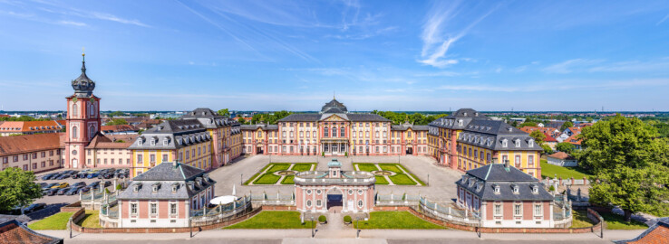 Schloss Bruchsal - Zentrum der Schlossanlage - Von Außen - Von Osten - Blick von der Plattform des Kanzleibaus nach unten auf die Dreiflügelanlage; in der Mitte der Hauptbau / das Corps de Logis, links der Mitte der südliche Verbindungsbau, links davon der Kirchenflügel / die Hofkirche / St. Damian und Hugo, links davon der Kirchturm, rechts der Mitte der nördliche Verbindungsbau, rechts davon der Kammerflügel / das Amtsgericht; vorne links das Hofzahlamt (heute Schlosscafé), vorne in der Mitte das Torwachtgebäude, vorne rechts das Hofkontrollamt; in der Mitte der Anlage der Ehrenhof (aufgenommen im Juni 2021, am späten Vormittag)