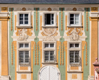 Schloss Bruchsal - Nördliche Orangerie / Nebendienstdienerbau - Von außen - Von Süden - Blick vom Gartenbalkon auf die Südfassade der nördlichen Orangerie mit der illusionistischen Fassadenmalerei (ursprünglich gemalt von Giovanni Francesco Marchini, zwischen 1732 und 1737, restauriert von Josef Mariano Kitschker, zwischen 1900 und 1910, erneut restauriert von Klaus Siller (Fassadenmalerei) und  Walter Maschke (Allegorien), 1976) (aufgenommen im Juli 2021, am späten Nachmittag)