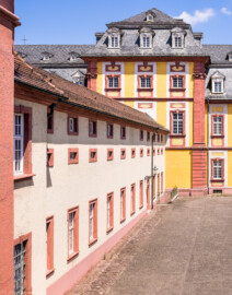 Schloss Bruchsal - Finsterer Gang / Hofkirche - Von Außen - Von Süden - Blick aus dem "Kleinen Saal" im 1. Obergeschoss des Seminarbaus nach Norden auf den Finsteren Gang / Verbindungsgang (links) sowie auf den Mittelrisalit der Hofkirche / St. Damian und Hugo (hinten) (aufgenommen im Juli 2021, am frühen Nachmittag)