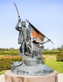 Waghäusel - Eremitage - Von Außen - Von Süden - Blick auf das Freiheitsdenkmal zum Gedenken an die Schlacht bei Waghäusel 1849 (Senkrechte Inschrift: "Den Wegbereitern der Demokratie - 21. Juni 1999"; schräge Inschrift: "In dankbarer Erinnerung an den Einsatz vieler Männer und Frauen für Freiheit und Menschenrechte in der Badischen Revolution von 1848-1849 wurde dieses Denkmal von der Stadt Waghäusel zum 150. Jahrestag der Schlacht von Waghäusel und des Gefechtes von Wiesental errichtet"; Inschrift in der Fahne: "Fond. Veneta d'Arte Verona Italia") (Skulptur aus Bronze, geschaffen 1999 von dem Speyerer Künstler Franz-Werner Müller-Steinfurt (1952-2017)) (aufgenommen im September 2021, am Nachmittag)