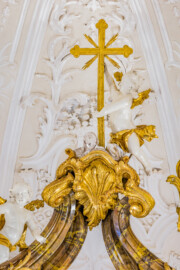 Bruchsal - Peterskirche / St. Peter - Hochaltar, ganz oben, Mitte - Blick von der Orgelempore auf das Kreuz ganz oben und auf den obersten Putto des Hochaltars (Altar entworfen von Balthasar Neumann und Johann Wolfgang van der Auwera, vor 1747) (aufgenommen im März 2022, am frühen Nachmittag, nach der Renovierung)