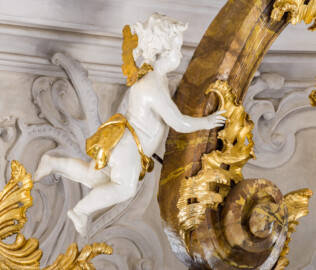 Bruchsal - Peterskirche / St. Peter - Hochaltar, oben, links - Blick von der Orgelempore auf den oberen linken Putto des Hochaltars (oberhalb des Architravs) (Altar entworfen von Balthasar Neumann und Johann Wolfgang van der Auwera, vor 1747) (aufgenommen im März 2022, am Nachmittag, nach der Renovierung) Bruchsal - Peterskirche / St. Peter - Hochaltar, oben, links - Blick von der Orgelempore auf den oberen linken Putto des Hochaltars (oberhalb des Architravs) (Altar entworfen von Balthasar Neumann und Johann Wolfgang van der Auwera, vor 1747) (aufgenommen im März 2022, am Nachmittag, nach der Renovierung)