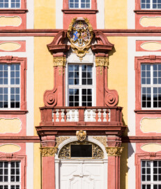Schloss Bruchsal - Kammerflügel - Von Süden / Hofseite - Mittelrisalit - Blick vom Ehrenhof auf das Hauptportal, den Balkon sowie das Prunkwappen von Kardinal Damian Hugo Philipp von Schönborn-Buchheim - Hinter dem Fenster des Balkons befindet sich der Kammermusiksaal - Wappenschild links: je zwei Felder für das Hochstift Speyer und die Propstei Weißenburg, Wappenschild unten: Deutschordens-Würden (Landkomtur des Deutschen Ordens in Alden-Biesen (oben) und  Marburg (unten)), Wappenschild rechts: Familien- / Stammwappen von Schönborn (aufgenommen im März 2022, am Nachmittag)