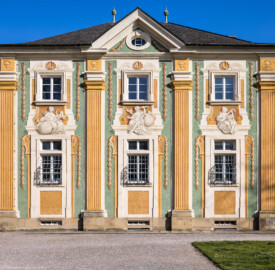 Schloss Bruchsal - Südliche Orangerie / Kavaliersbau - Von Außen / Von Westen - Blick von Terrasse auf die nördliche Westfassade der Orangerie (Fassade der Orangerie ursprünglich gemalt von Giovanni Francesco Marchini, zwischen 1732 und 1737, restauriert von Josef Mariano Kitschker, zwischen 1900 und 1910, erneut restauriert von Klaus Siller (Fassadenmalerei) und  Walter Maschke (Allegorien), 1976) (aufgenommen im März 2022, am späten Nachmittag)