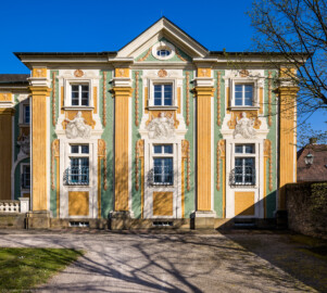 Schloss Bruchsal - Südliche Orangerie / Kavaliersbau - Von Außen / Von Westen - Blick von der südlichsten Ost-West-Wegeachse auf die südliche Westfassade der Orangerie (Fassade der Orangerie ursprünglich gemalt von Giovanni Francesco Marchini, zwischen 1732 und 1737, restauriert von Josef Mariano Kitschker, zwischen 1900 und 1910, erneut restauriert von Klaus Siller (Fassadenmalerei) und  Walter Maschke (Allegorien), 1976) (aufgenommen im März 2022, am späten Nachmittag)