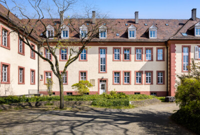 Schloss Bruchsal - Landhospital / Seminarbau - Innenhof, Westwand, nördlich des Durchgangs - Von Außen - Von Westen - Blick auf die Westwand des Seminarbaus, links die Südwand, rechts der Mittelrisalit mit dem Durchgang zur Schönbornstraße; links der Tür das Portraitrelief von Johann Peter Frank (1745-1821) (aufgenommen im April 2022, am Nachmittag)