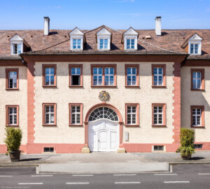 Schloss Bruchsal - Landhospital / Seminarbau - Mittelrisalit des Mitteltrakts - Von Außen - Von Osten - Blick auf den Mittelrisalit, den Durchgang nach Westen sowie auf das Landeswappen des Fürstbistums Speyer zur Regierungszeit des Fürstbischofs Kardinal Damian Hugo Philipp von Schönborn-Buchheim (aufgenommen im April 2022, am frühen Nachmittag)