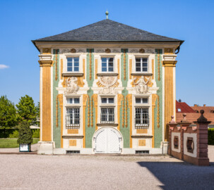 Schloss Bruchsal - Nördliche Orangerie / Nebendienstdienerbau - Von außen - Von Süden - Blick auf die Südfassade der nördlichen Orangerie mit der illusionistischen Fassadenmalerei (ursprünglich gemalt von Giovanni Francesco Marchini, 1732, restauriert von Josef Mariano Kitschker, zwischen 1900 und 1910, erneut restauriert von Klaus Siller (Fassadenmalerei) und  Walter Maschke (Allegorien), 1976) (aufgenommen im April 2022, am Nachmittag)
