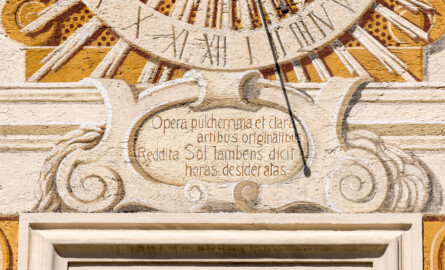 Schloss Bruchsal - Nördliche Orangerie / Nebendienstdienerbau - Von außen - Von Süden - Südfassade - Spruch unterhalb der Sonnenuhr - Inschrift: "Opera pulcherrima et clara / artibus originalibus / Reddita Sol lambens dicit / horas desideratas" / "Während die Sonne auf die schönen und herrlichen Bauwerke, die mit originaler Kunst aufgeführt sind, ihre Strahlen wirft, zeigt sie ihre Stunden an", übersetzt von Bruno Schallbach (ursprünglich gemalt von Giovanni Francesco Marchini, zwischen 1732 und 1737, restauriert von Josef Mariano Kitschker, zwischen 1900 und 1910, erneut restauriert von Klaus Siller (Fassadenmalerei) und  Walter Maschke (Allegorien), 1976) (aufgenommen im April 2022, am Nachmittag)