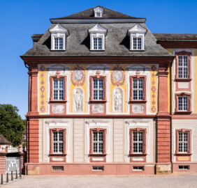 Schloss Bruchsal - Kammerflügel / Amtsgericht - Von Westen - Blick vom Nebendienstdienerbau auf die Westfassade des Kammerflügels mit den Figuren von Paris (Sohn des trojanischen Königs Priamos) und Venus (römische Göttin der Liebe und der Schönheit) (gemalt von Walter Maschke), fünf Landschaftgrisaillen und der illusionistischen Fassadenmalerei (ursprünglich gemalt von Giovanni Francesco Marchini, zwischen 1732 und 1737) (aufgenommen im Juli 2022, am späten Nachmittag)