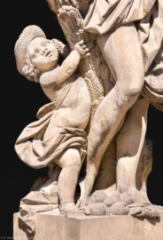 Schloss Bruchsal - Garten - Hauptachse, Nordseite, 1. Skulptur von Osten - Kindliche Figur links unten der Skulptur "Der Sommer" aus dem Skulpturenensemble "Vier Jahreszeiten" (Kopie; Original gehauen 1759 von Joachim Günther, befindet sich im Harvard Art Museum / Fogg Museum) (aufgenommen im Juli 2022, am Nachmittag)