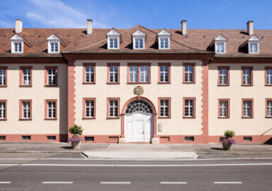 Schloss Bruchsal - Großer Dienerbau / Finanzamt - Mitteltrakt / Mittelrisalit - Von Außen - Von Westen - Blick auf die Westfassade und den Mittelrisalit des Großer Dienerbaus; über dem Durchgang das Landeswappen des Fürstbistums Speyer zur Regierungszeit des Fürstbischofs Kardinal Damian Hugo Philipp von Schönborn-Buchheim (aufgenommen im Juli 2022, am späten Nachmittag)