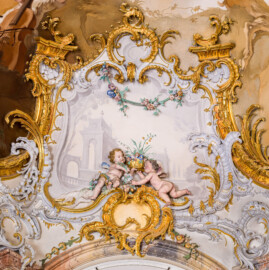 Schloss Bruchsal - Hauptbau / Corps de Logis - Beletage - Marmorsaal / Kaisersaal - Nordostecke, oben, Stuckfeld - Von Südwesten - Blick auf zwei Putti (als allegorische Darstellung des Frühlings) sowie auf umgebende Rokoko-Stuckaturen (Stuckaturen ursprünglich von Johann Michael Feichtmayr) (aufgenommen im August 2022, um die Mittagszeit)
