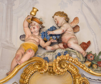 Schloss Bruchsal - Hauptbau / Corps de Logis - Beletage - Marmorsaal / Kaisersaal - Südwestecke, oben, Stuckfeld - Von Nordosten - Blick auf zwei Putti als allegorische Darstellung des Herbsts (Stuckaturen ursprünglich von Johann Michael Feichtmayr) (aufgenommen im August 2022, am frühen Nachmittag)