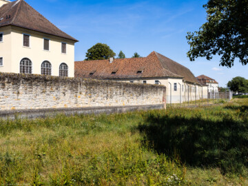 Bad Schönborn / Bad Mingolsheim - Schloss Kislau - Küchenbau / Südlicher Remisenanbau / Östliche Remisen und Pferdeställe - Von Südosten - Blick vom Landgraben auf den Küchenbau (links), den südlichen Remisenanbau (Mitte), den östlichen Bau für Remisen und Pferdeställe (rechts) sowie auf die Umgebungsmauer; ganz rechts im Hintergrund ein neuerer Bau (aufgenommen im August 2022, am späten Vormittag)