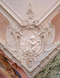 Bad Schönborn / Bad Mingolsheim - Schloss Kislau - Hauptbau - Von Innen - Schlafzimmer - Decke, Nordwestecke - Blick nach oben auf die Stuckverzierung der nordwestlichen Ecke der Decke des Schlafzimmers; im Zentrum ein spielendes Kind und allegorische Andeutungen der Jahreszeit Winter und des Elements Feuer; Stuckverzierungen erstellt vor 1770 von Joachim Günther (1720-1789)  (aufgenommen im September 2022, am Vormittag)