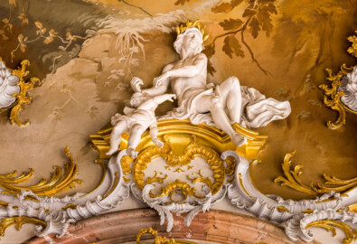 Schloss Bruchsal - Hauptbau / Corps de Logis - Beletage - Marmorsaal / Kaisersaal - Stuckaturzone, oberhalb des Durchgangs zum Gelben Zimmer - Blick auf Figuren, einen Mann mit Lorbeerkranz und einen Putto (Deckenbild ursprünglich von Johannes Zick, rekonstruiert von Wolfram Köberl, 1969 bis 1973; Stuckaturen ursprünglich von Johann Michael Feichtmayr) (aufgenommen im März 2023, am Nachmittag)